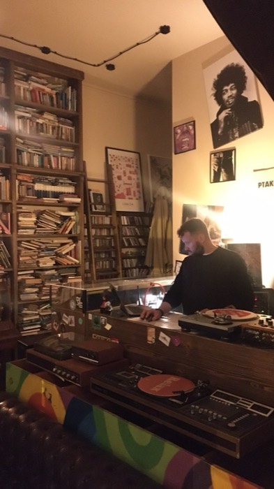 DJ spinning vinyl in cozy book-lined bar, Hendrix poster, Lublin print