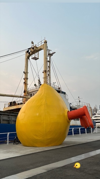 Giant yellow duck-butt sculpture diving on pier beside cargo ship