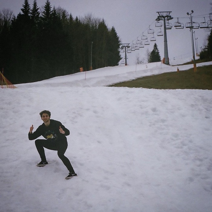 Joshua: 35mm film selfie crouching on snowy ski slope, chairlifts behind