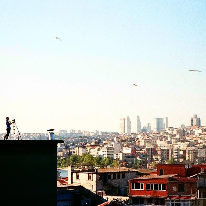 35mm Istanbul rooftop panorama, silhouetted man filming on tripod, seagulls overhead