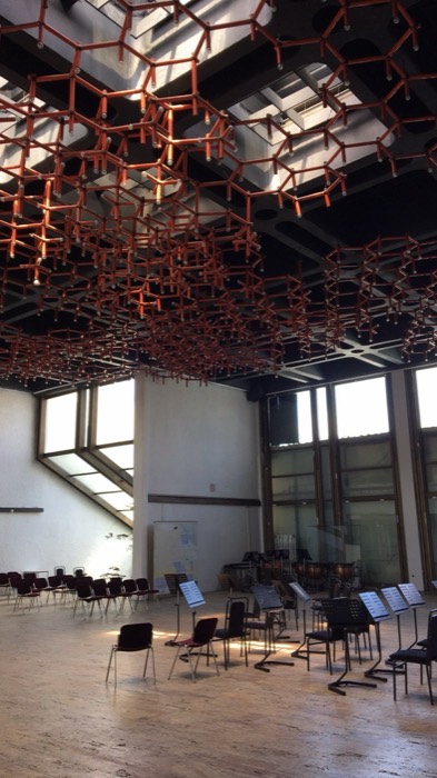 Brutalist concert hall interior, red hexagonal ceiling sculpture above empty orchestra chairs
