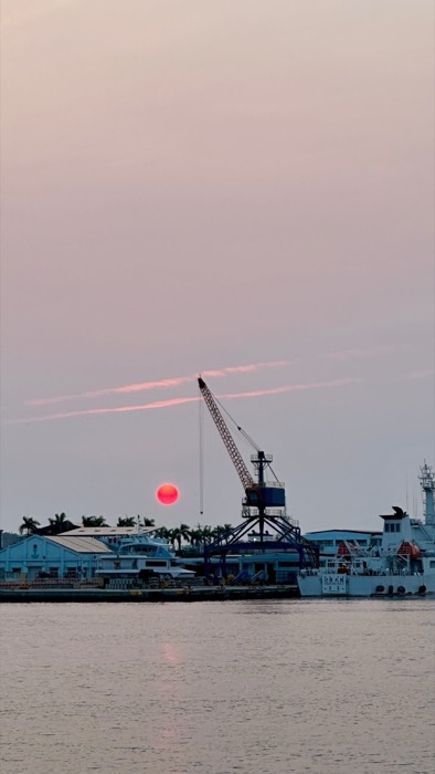 Blood-red sun setting behind harbor crane and coast guard ship