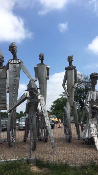 Quirky giant tin-man sculptures behind fence by a parking lot