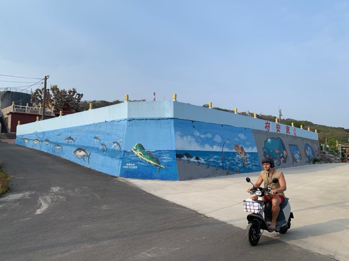 Scooter rider passing a long blue fish mural along a coastal road
