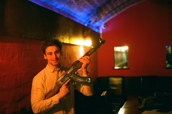 Joshua: poses holding replica AK-47 in red-lit cellar bar.