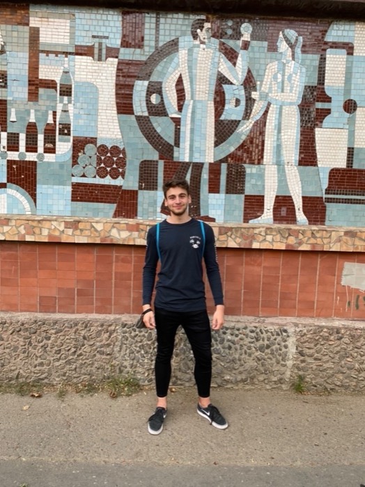 Joshua: full-length standing portrait smiling before colourful Soviet mosaic mural.
