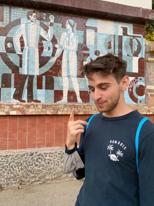 Joshua: close portrait pointing up at himself before Soviet-era mosaic mural.