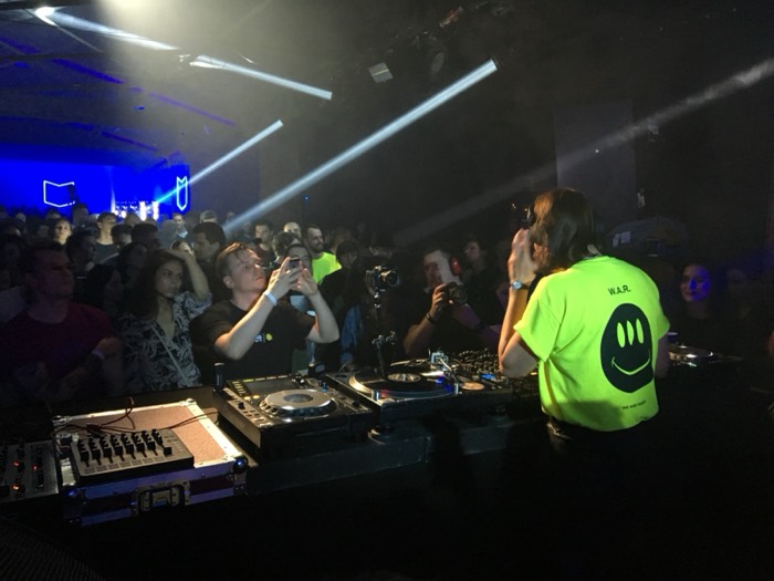 DJ in neon WAR shirt, crowd filming under light beams