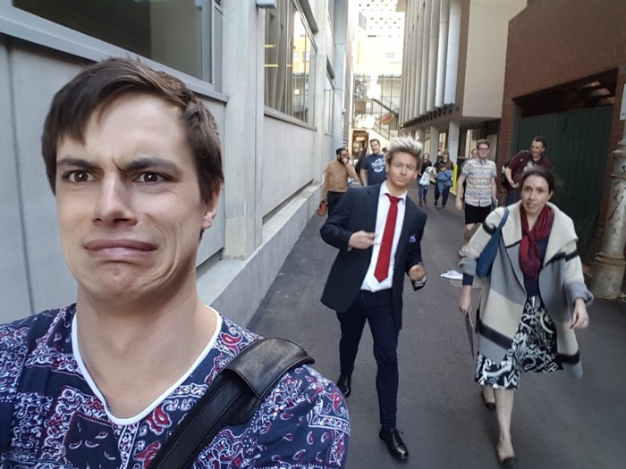 Joshua: grimacing selfie in laneway with people walking behind