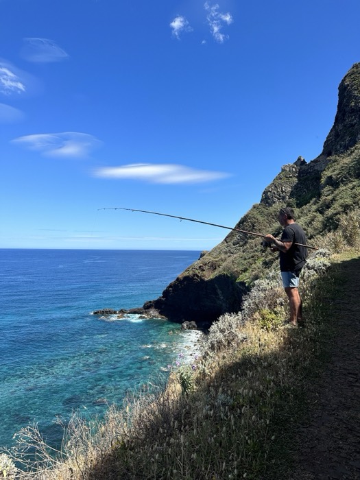 Man fishing from clifftop trail above blue ocean, coastal hike