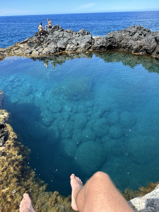 Joshua: legs dangling over clear volcanic tidal pool, two swimmers