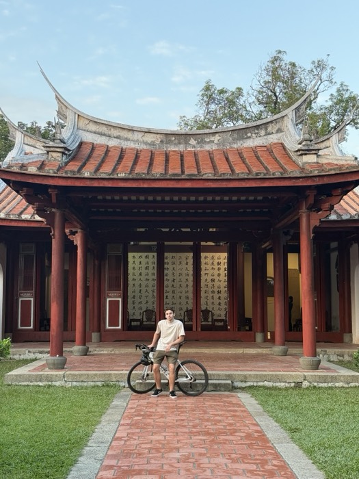 Joshua: leans on bike before grand red Confucian temple with curved roof