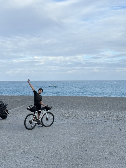 Joshua: celebrating arm raised beside loaded bike on pebble beach, Taiwan