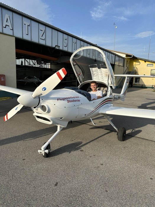 Joshua: smiling in cockpit of Diamond DA20 plane, canopy open, hangar behind