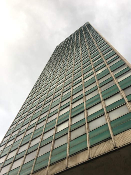 Looking up at green modernist glass tower against grey sky.