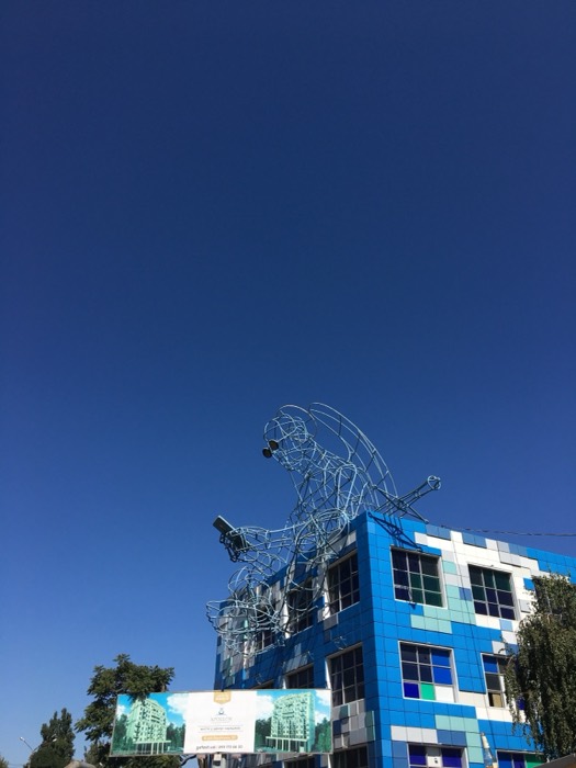 Wire-frame angel sculpture atop blue-tiled building, deep blue sky.