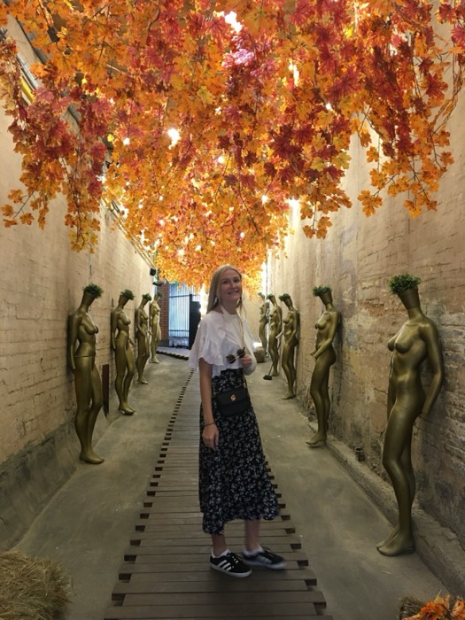 Friend in golden-mannequin alley under autumn-leaf canopy art installation.