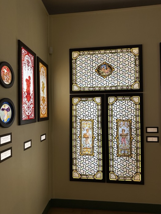 Stained-glass panels in dim museum corner, glowing colour against khaki walls