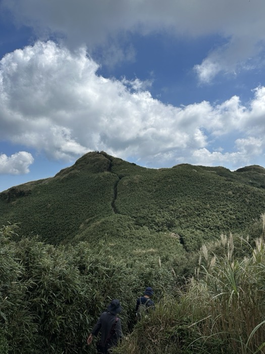 Two hikers ascending grassy ridge trail toward cloudy peak