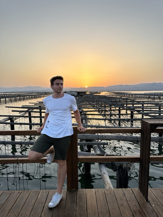 Joshua: leaning on rail at oyster-farm pier, golden sunset, white tee