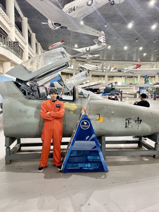 Joshua: posing in orange flight suit beside fighter jet in aviation museum