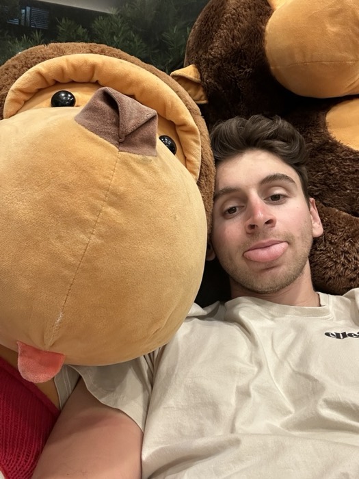 Joshua: tongue-out selfie cuddled beside giant brown monkey plush toys.
