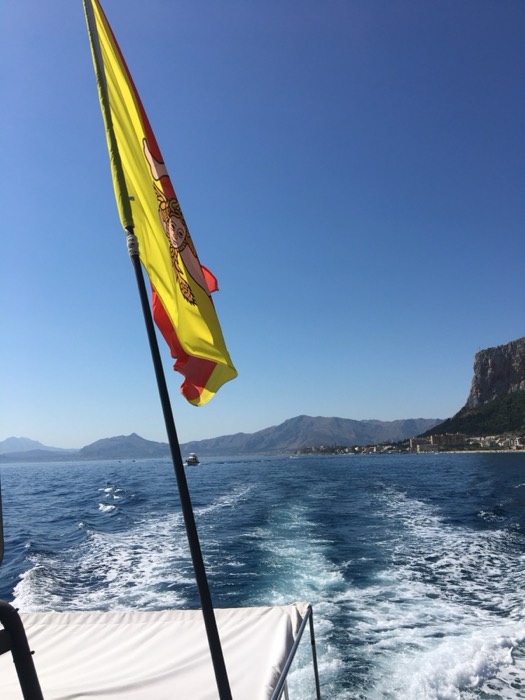 Sicilian flag flying off boat stern, coastal cliffs and wake behind