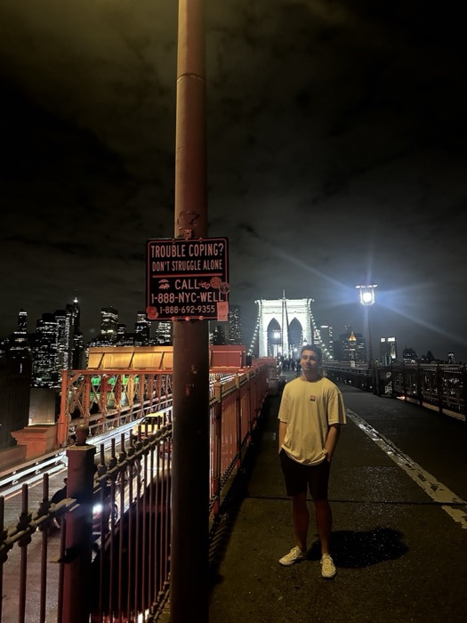 Joshua: on Brooklyn Bridge at night with lit Manhattan skyline, Trouble Coping sign.