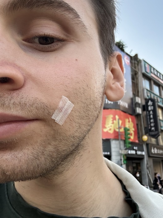 Joshua: close selfie with surgical tape on cheek, Taiwan street signage behind