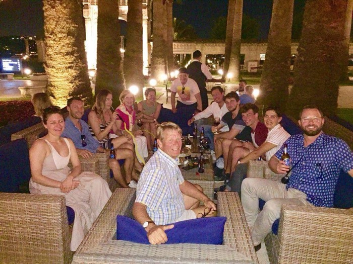 Group of friends relaxing on outdoor lounges among lit palm trees, evening drinks