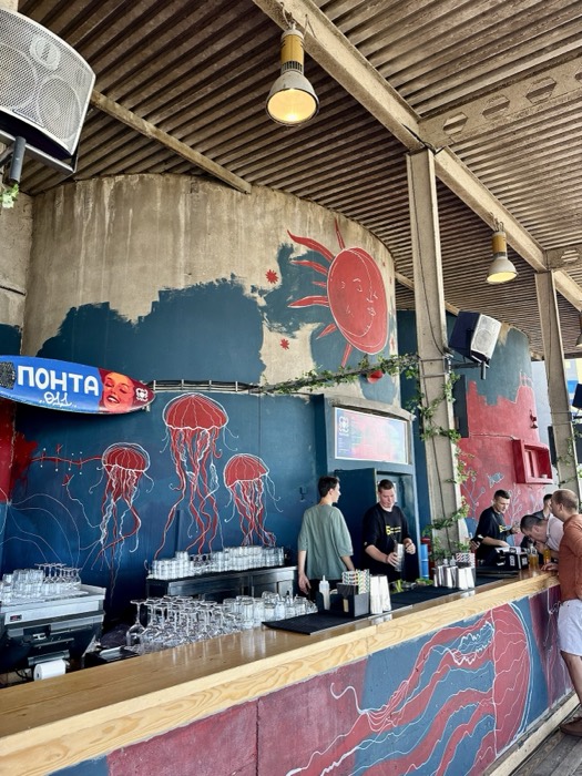 Riverside bar with bold red jellyfish and sun mural, bartenders working