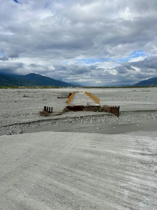 Washed-out footbridge across broad grey riverbed, mountains beyond