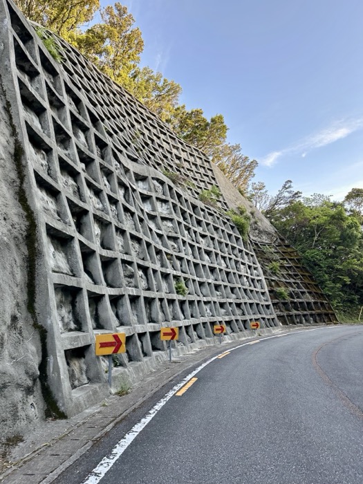 Massive cribbed concrete slope wall with orange arrow signs, mountain road.