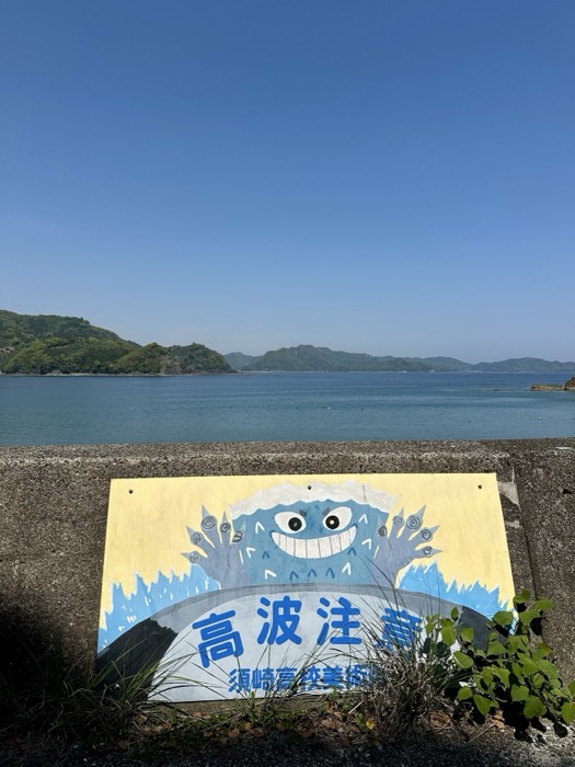 Grinning wave-monster mural high-wave warning sign above calm island bay.