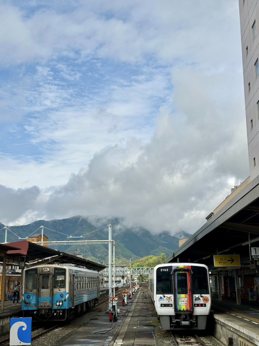 Station scene: local train and rainbow Anpanman train below cloudy mountains