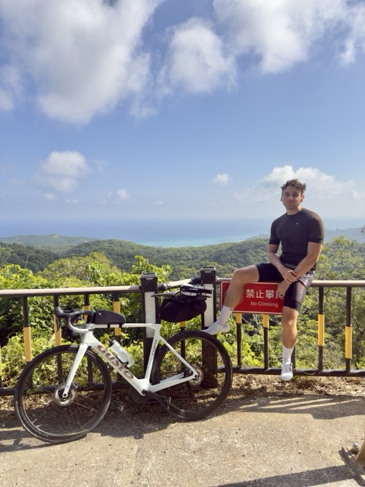 Joshua: cyclist on No Climbing rail above turquoise bay, bike below