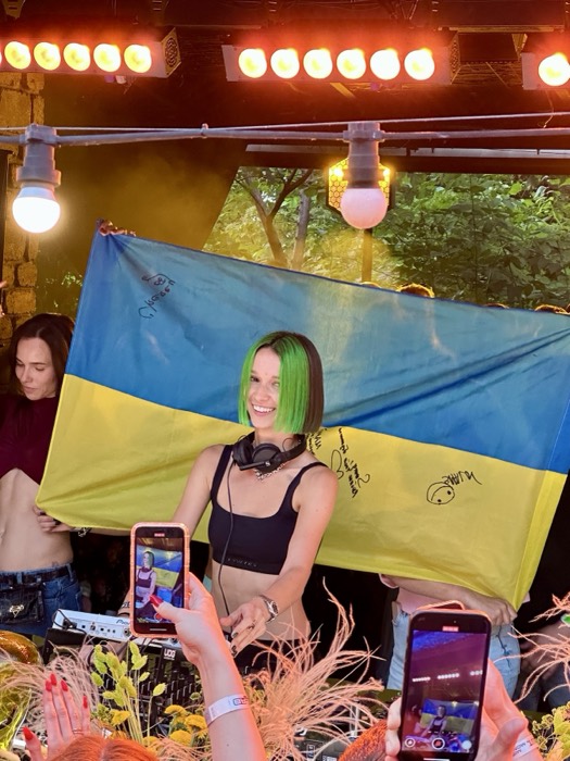 Green-haired DJ smiling behind signed Ukrainian flag, crowd phones up