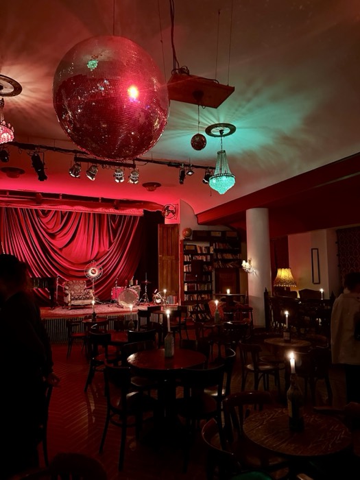 Moody red-lit bar: giant disco ball, draped stage, candlelit tables.