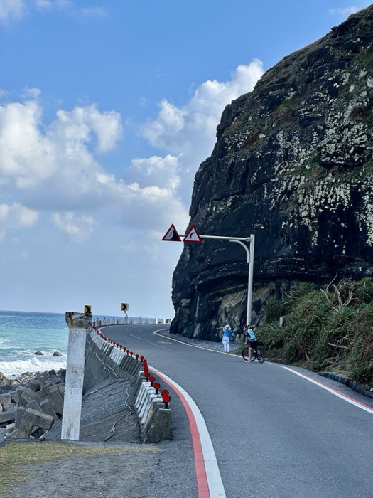 Dramatic black-cliff coastal road, cyclist and warning signs, turquoise sea.