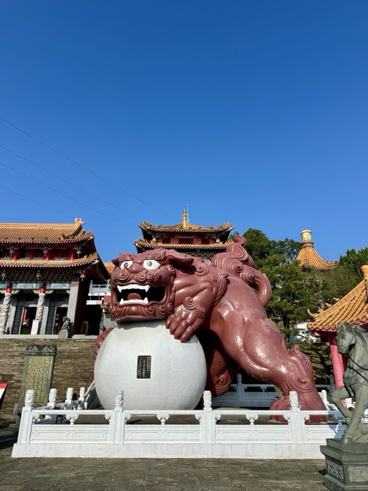 Bold red guardian lion sculpture on ball before golden temple roofs