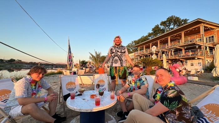 Friends in Hawaiian shirts and leis at sunset riverside beach bar.