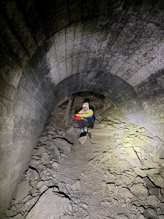 Headlamp-lit person crouched inside collapsed arched railway tunnel full of rubble