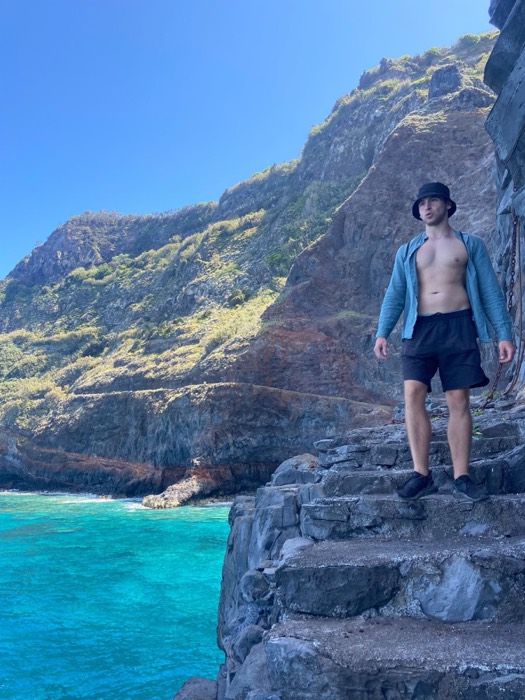 Joshua: shirtless in bucket hat on volcanic rock above turquoise sea